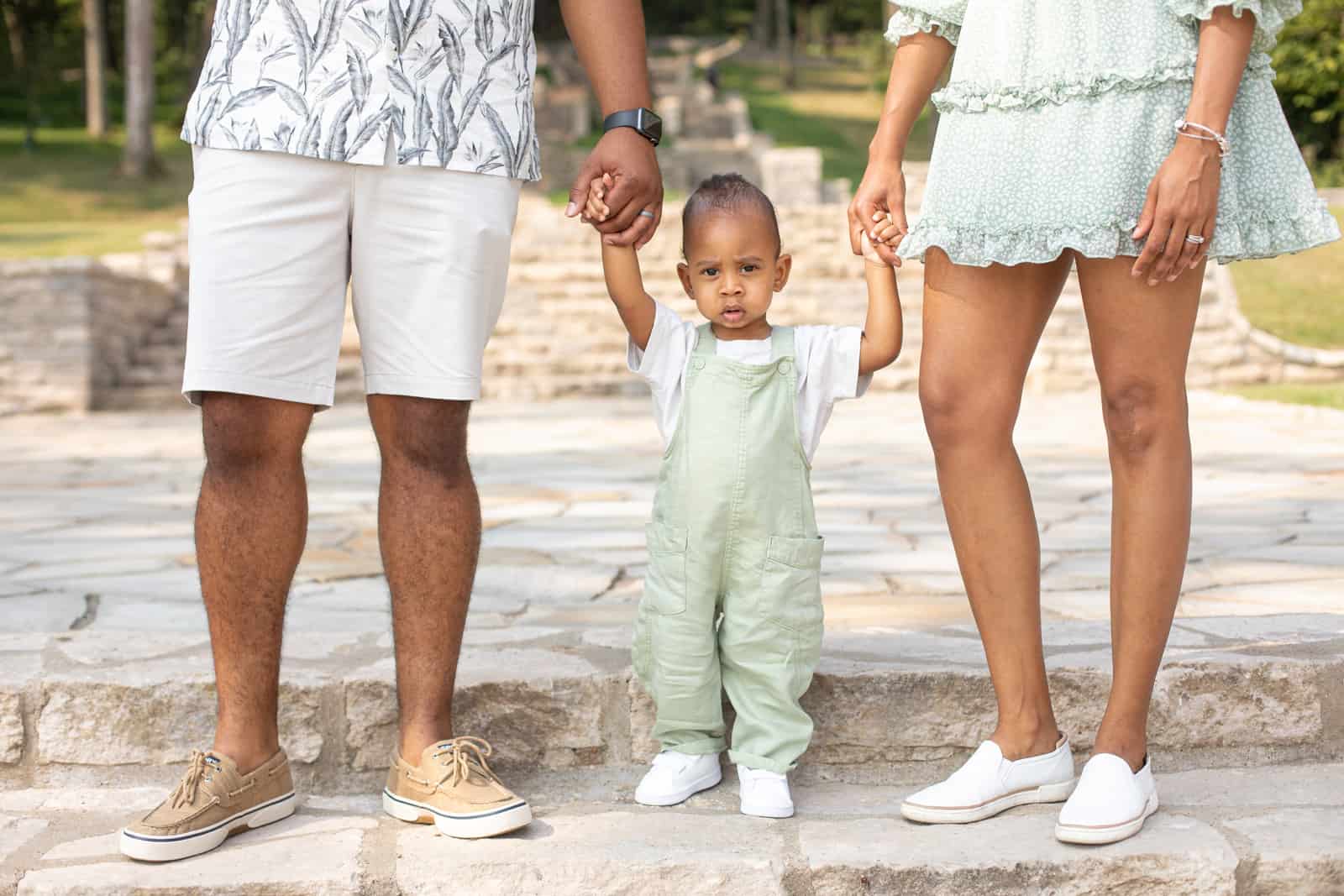 Black couple hold the hands of their one year old baby at the park.